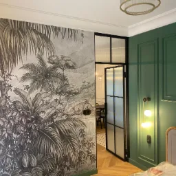 Stylowe wnętrze z dekoracyjną tapetą w motywy roślinne i zieloną ścianą z eleganckimi panelami. Widoczna czarna, przeszklona rama drzwi i designerskie oświetlenie.