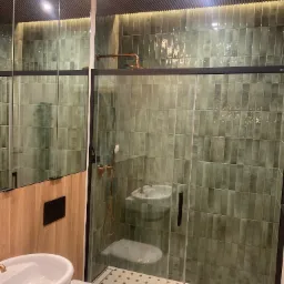 Nowoczesna łazienka z zielonymi płytkami typu 'cegiełka', kabiną prysznicową ze szkła hartowanego i armaturą w kolorze mosiądzu. Na podłodze wzorzysta mozaika.