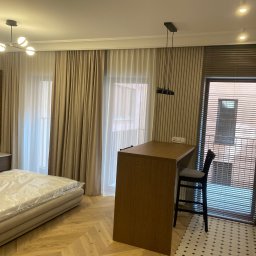 Piotr Krzeminski Pod Klucz - Nowoczesne wnętrze: łóżko z folią ochronną, drewniany parkiet w jodełkę, barek z krzesłem, żaluzje i dekoracyjne lampy sufitowe. Neutralna kolorystyka.