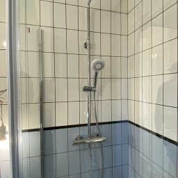 Piotr Krzeminski Pod Klucz - Nowoczesna kabina prysznicowa z chromowaną armaturą i biało-błękitnymi płytkami w kratkę. Widoczna szklana ściana kabiny i deszczownica.