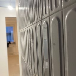 Piotr Krzeminski Pod Klucz - Ściana wyłożona błyszczącymi, szarymi płytkami z wypukłym, podłużnym wzorem. Widoczny fragment pomieszczenia w trakcie wykańczania, z surowym podłożem i otwartym przejściem.