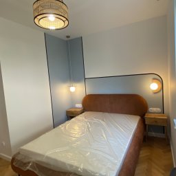 Wykończenia Pod Klucz Gdańsk - Piotr Krzeminski - Nowoczesna sypialnia z łóżkiem w odcieniach brązu, lampami nocnymi i parkietem w jodełkę. Materac zabezpieczony folią. Widoczne elementy dekoracyjne i oświetlenie.