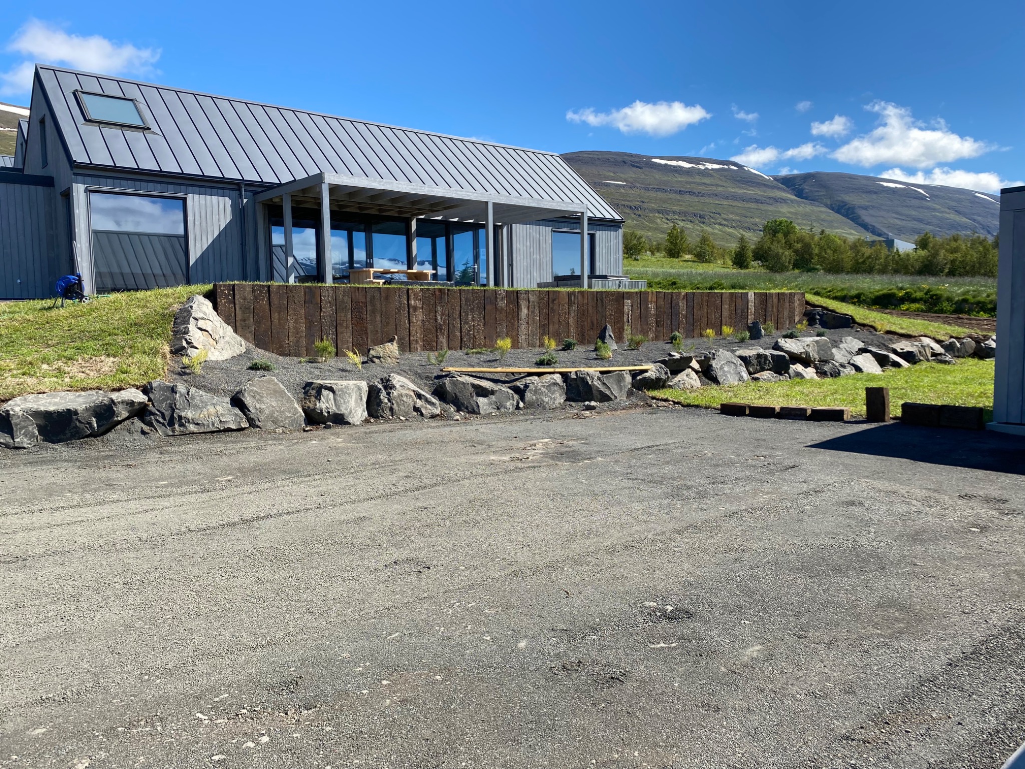 Nowoczesny dom z szarą elewacją i metalowym dachem, otoczony kamiennym i drewnianym murem oporowym oraz zielenią, na tle gór z resztkami śniegu.