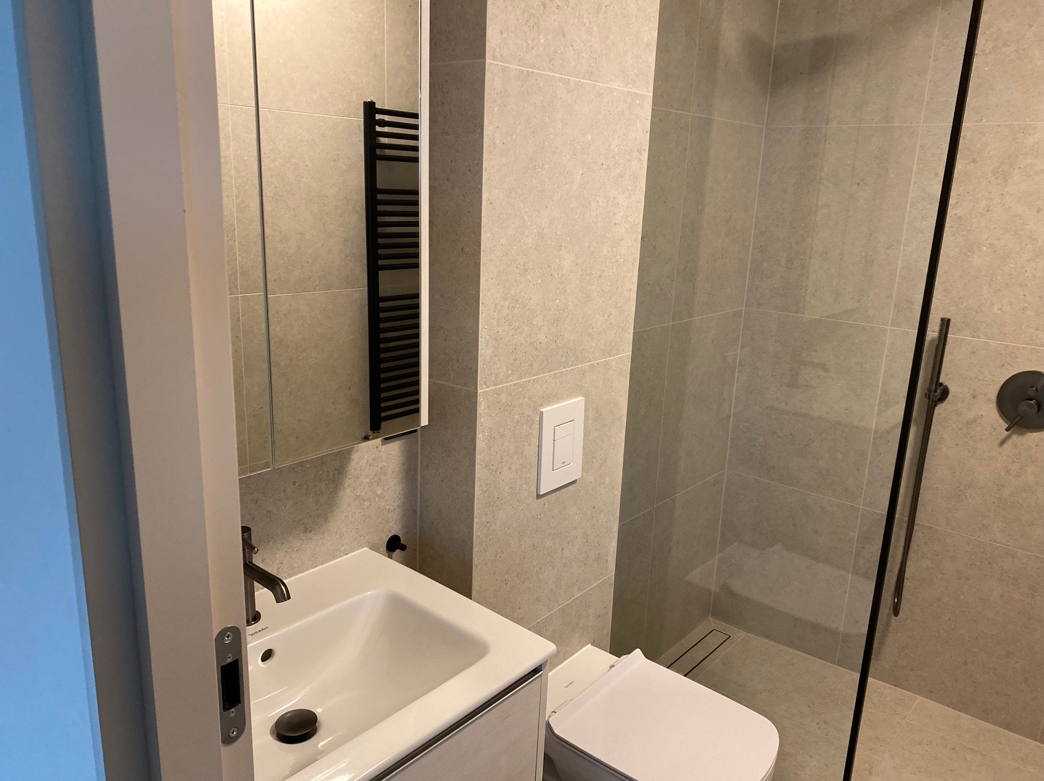 Nowoczesna łazienka z umywalką nablatową, podwieszaną toaletą i kabiną prysznicową typu walk-in, wykończona płytkami w odcieniach szarości, z czarnym grzejnikiem drabinkowym i minimalistyczną...