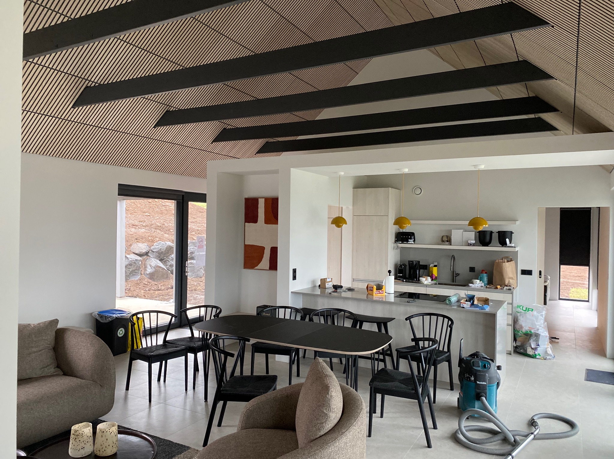 Nowoczesne wnętrze domu z drewnianym sufitem z listew, czarnymi belkami, szarymi ścianami, czarnymi krzesłami wokół czarnego stołu, widok na kuchnię z żółtymi lampami i odkurzaczem.