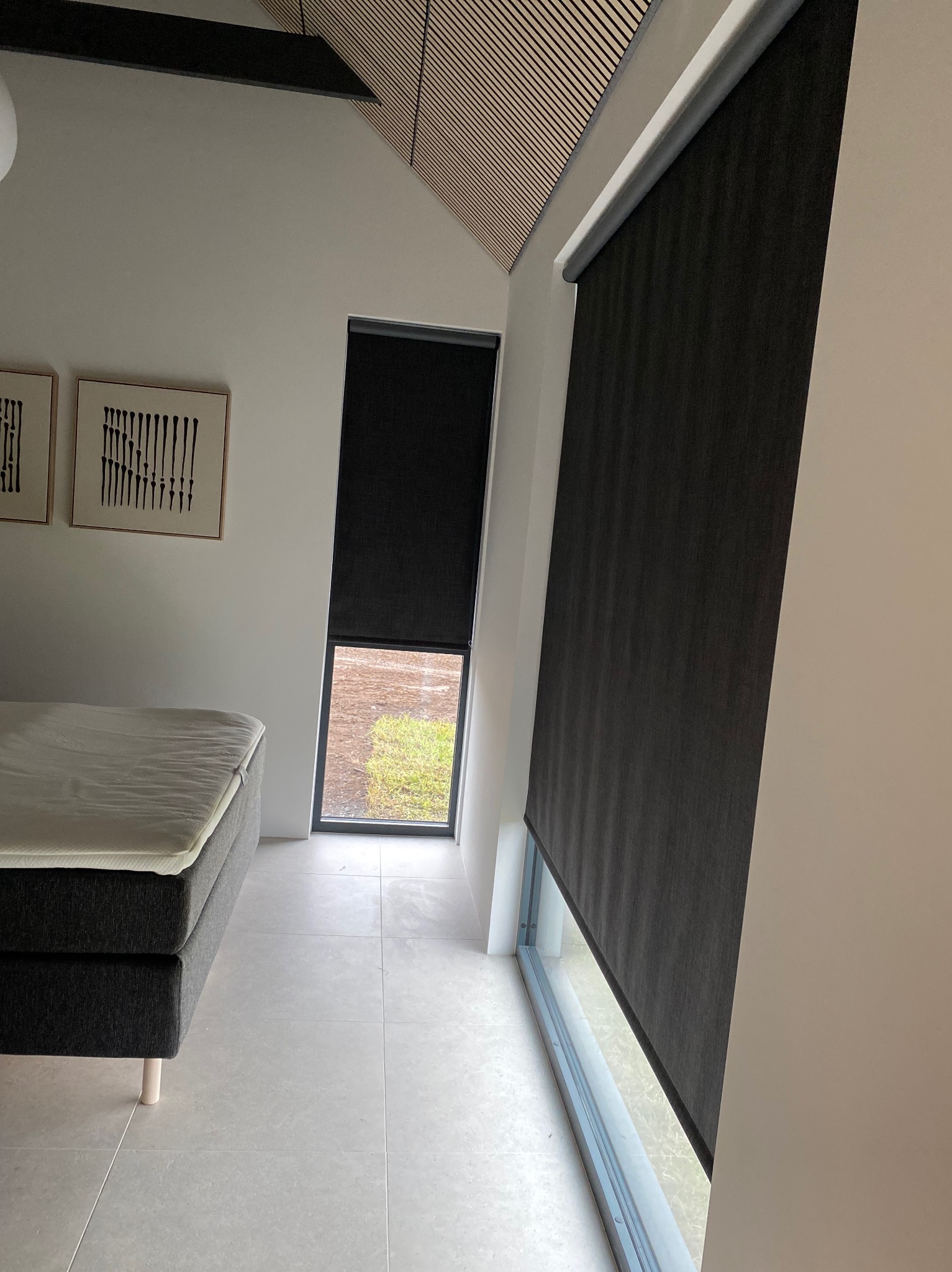 Nowoczesna sypialnia z drewnianym sufitem, szarym łóżkiem i zaciemniającymi roletami w oknach, widok na zewnątrz.