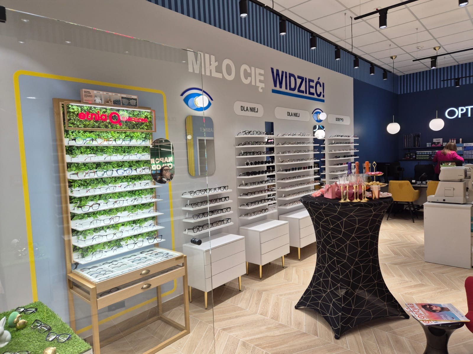 Wnętrze salonu optycznego z ekspozycją okularów, w tym regał z zielenią i okularami marki Ethnia Barcelona, oraz stolik z dekoracjami i napisem 'Miło Cię Widzieć!'.