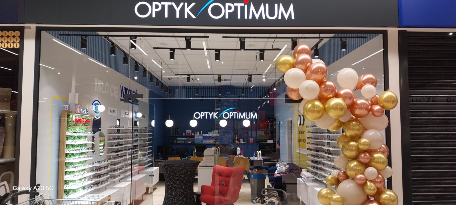 Wejście do salonu optycznego Optyk Optimum udekorowane girlandą z białych, złotych i różowych balonów. Wnętrze widoczne przez szklane drzwi z ekspozycją okularów i miejscami do siedzenia.