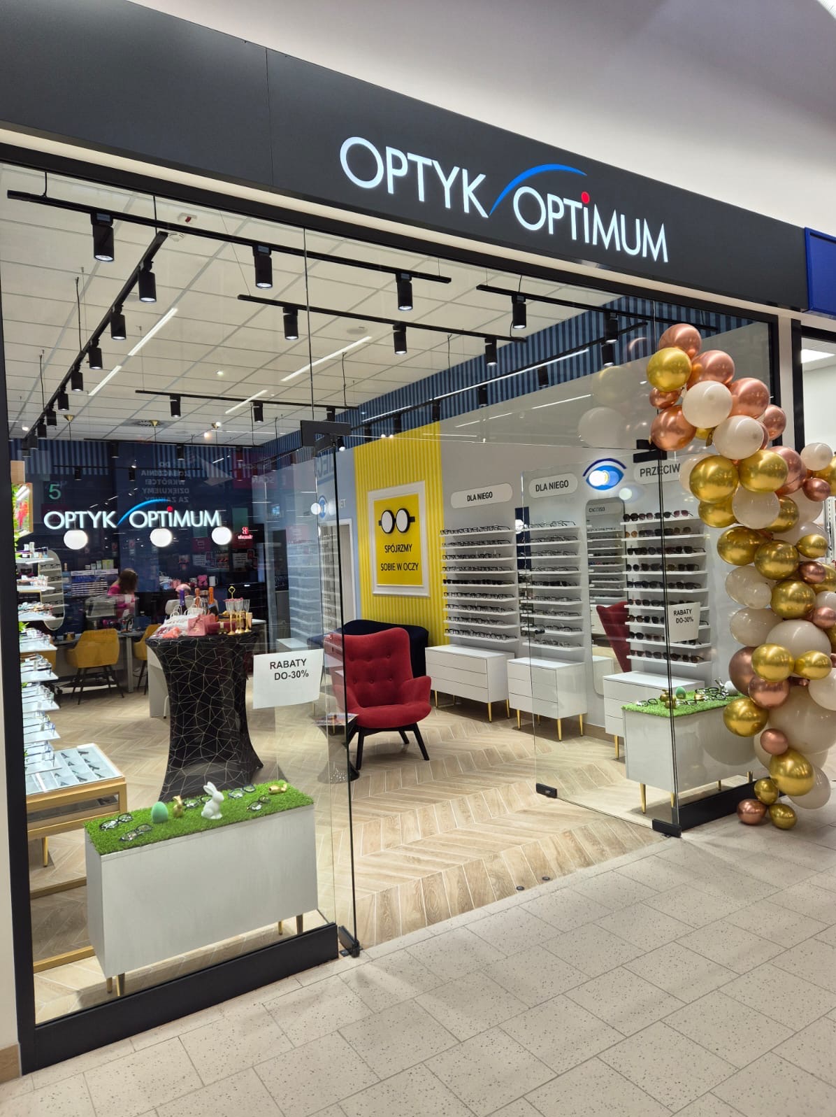 Fasada salonu optycznego Optyk Optimum z dekoracją balonową w odcieniach bieli, złota i różowego złota. Wnętrze widoczne przez szklane drzwi, z ekspozycją okularów, czerwonym fotelem i plakatem...