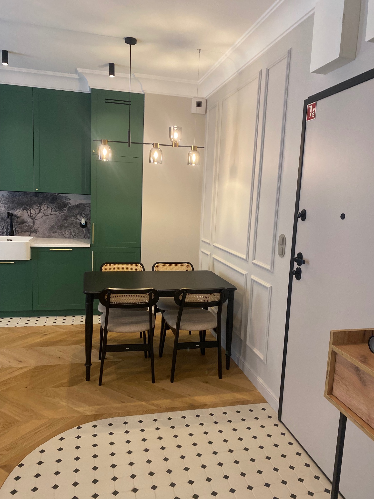 Nowoczesna jadalnia z zieloną zabudową kuchenną, drewnianym parkietem w jodełkę i białymi panelami ściennymi. Czarny stół i krzesła z rattanowymi oparciami.