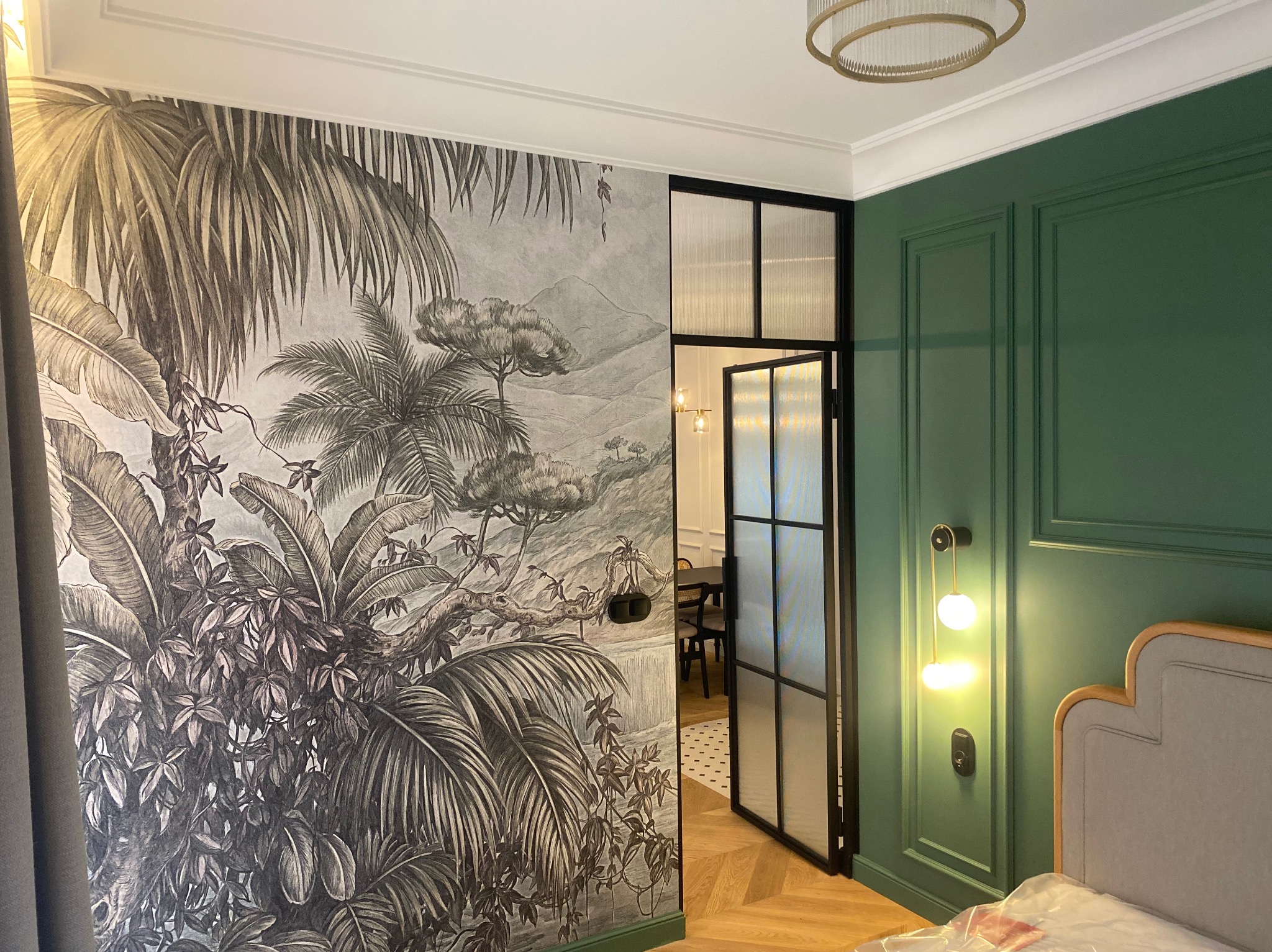 Stylowe wnętrze z dekoracyjną tapetą w motywy roślinne i zieloną ścianą z eleganckimi panelami. Widoczna czarna, przeszklona rama drzwi i designerskie oświetlenie.