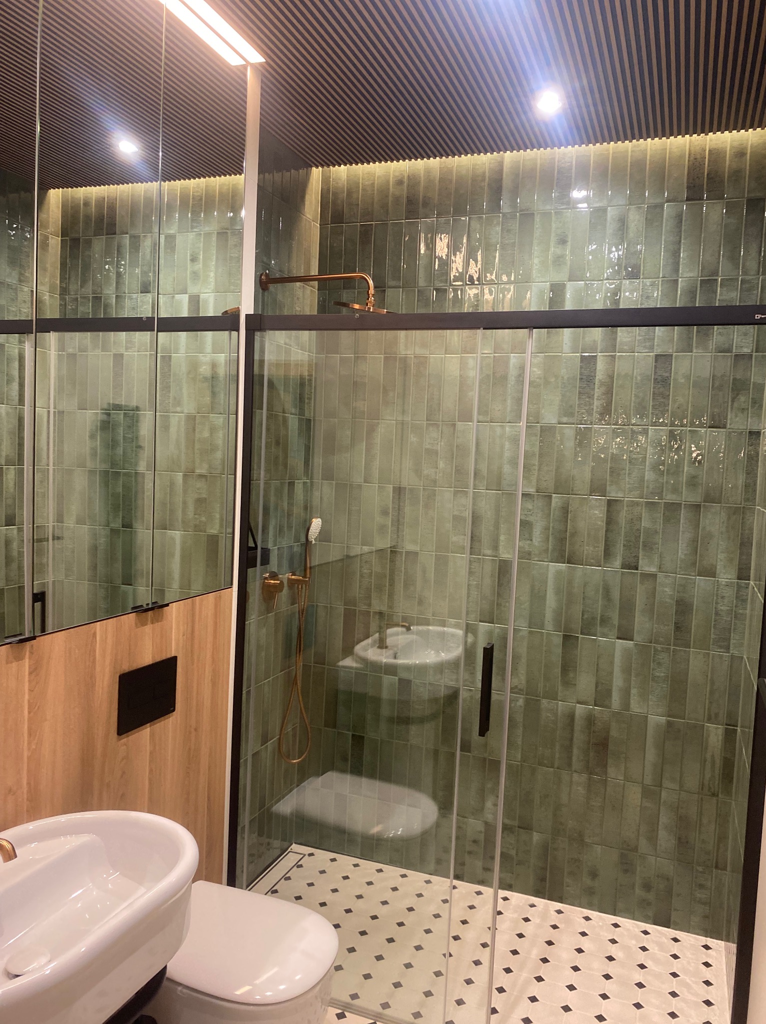 Nowoczesna łazienka z zielonymi płytkami typu 'cegiełka', kabiną prysznicową ze szkła hartowanego i armaturą w kolorze mosiądzu. Na podłodze wzorzysta mozaika.