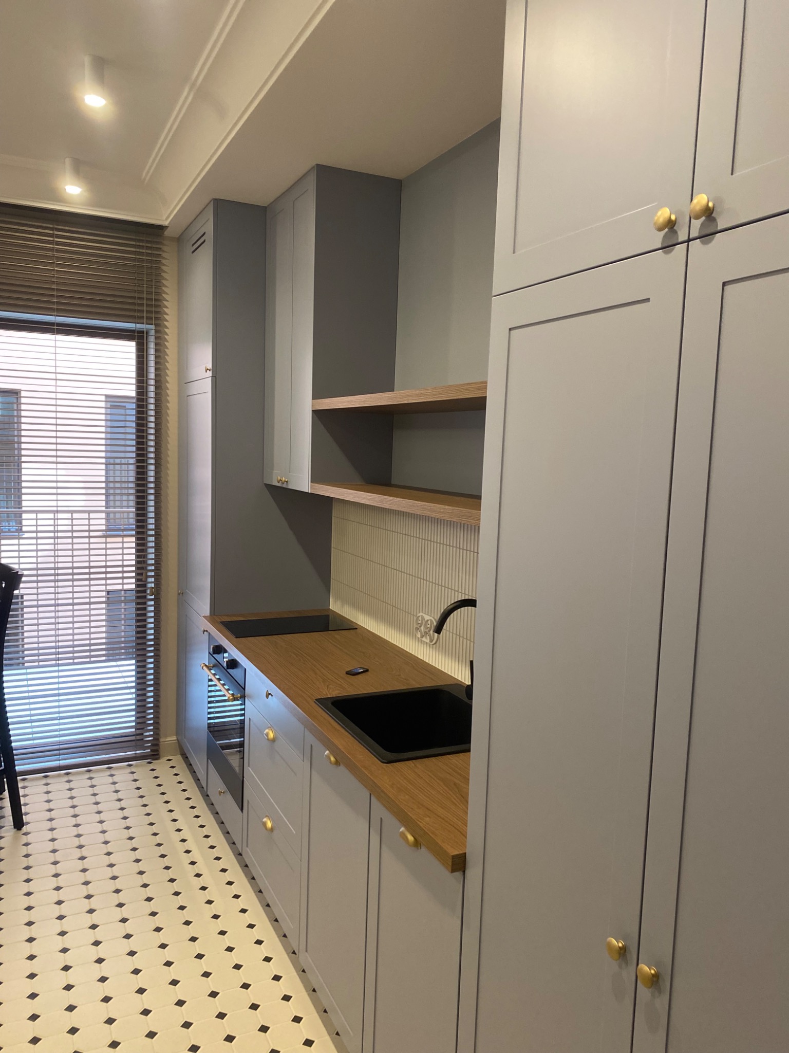 Szara kuchnia z drewnianym blatem i złotymi uchwytami, widok na okno z żaluzjami i wzorzystą podłogę. Nowoczesny design, detale wykończeniowe.