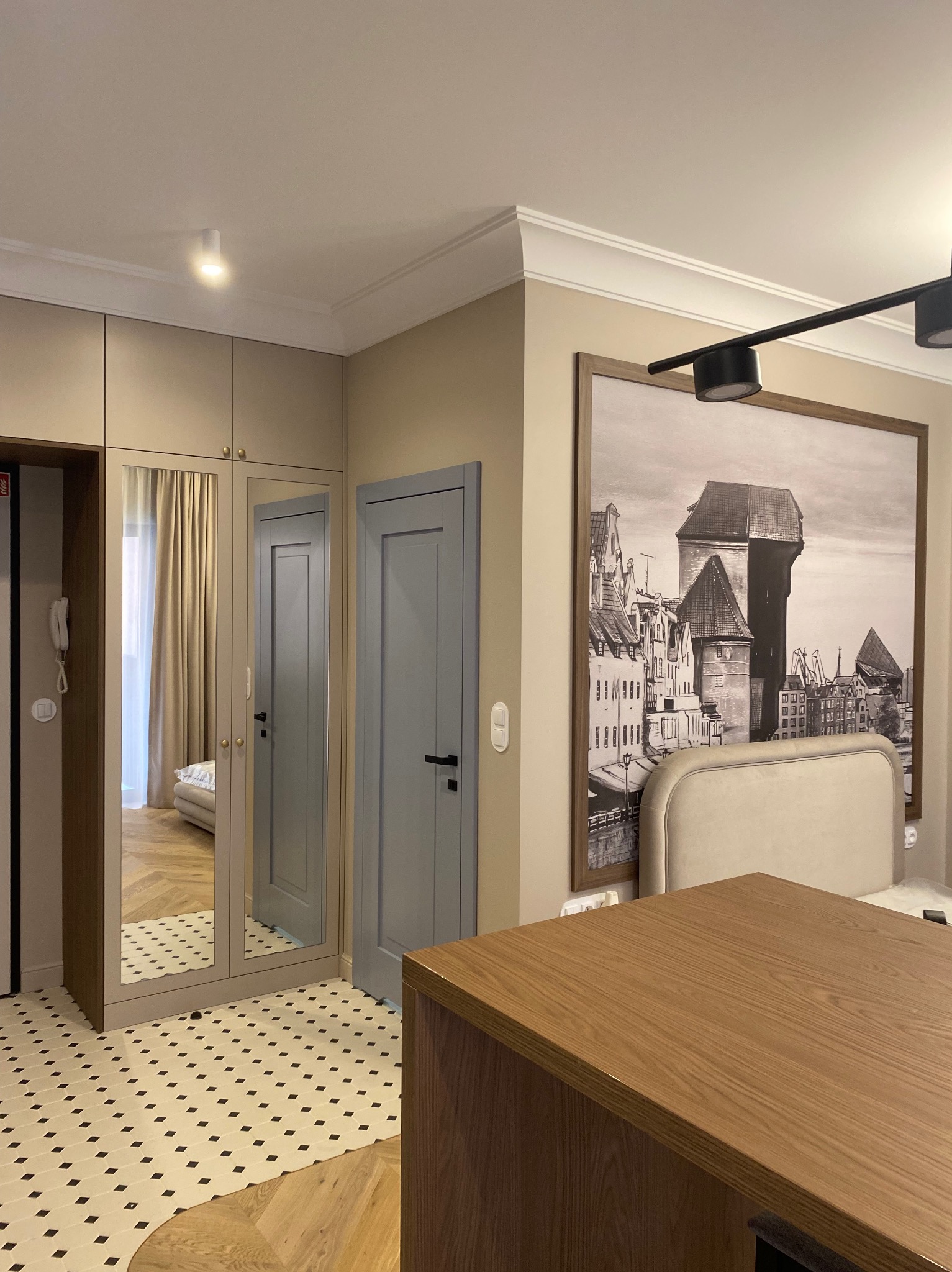 Nowoczesne wnętrze: szafa z lustrem, szara stolarka drzwiowa i mozaika na podłodze. Na ścianie duży obraz z motywem miasta. Całość w jasnej, stonowanej kolorystyce.