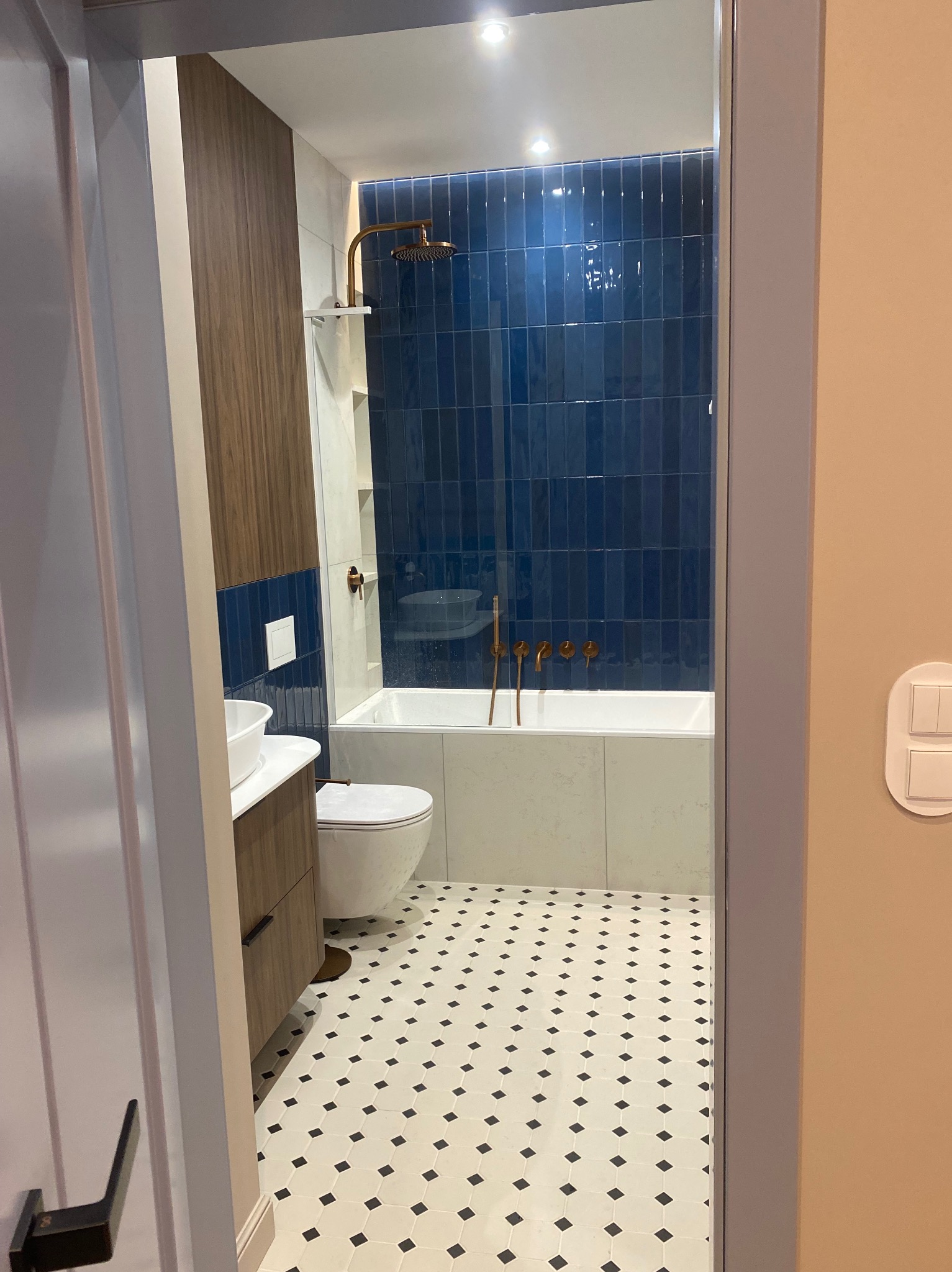 Nowoczesna łazienka z niebieskimi płytkami, złotą armaturą i wzorzystą podłogą, widziana zza uchylonych drzwi. Uwagę przykuwa oryginalny design i dbałość o detale.