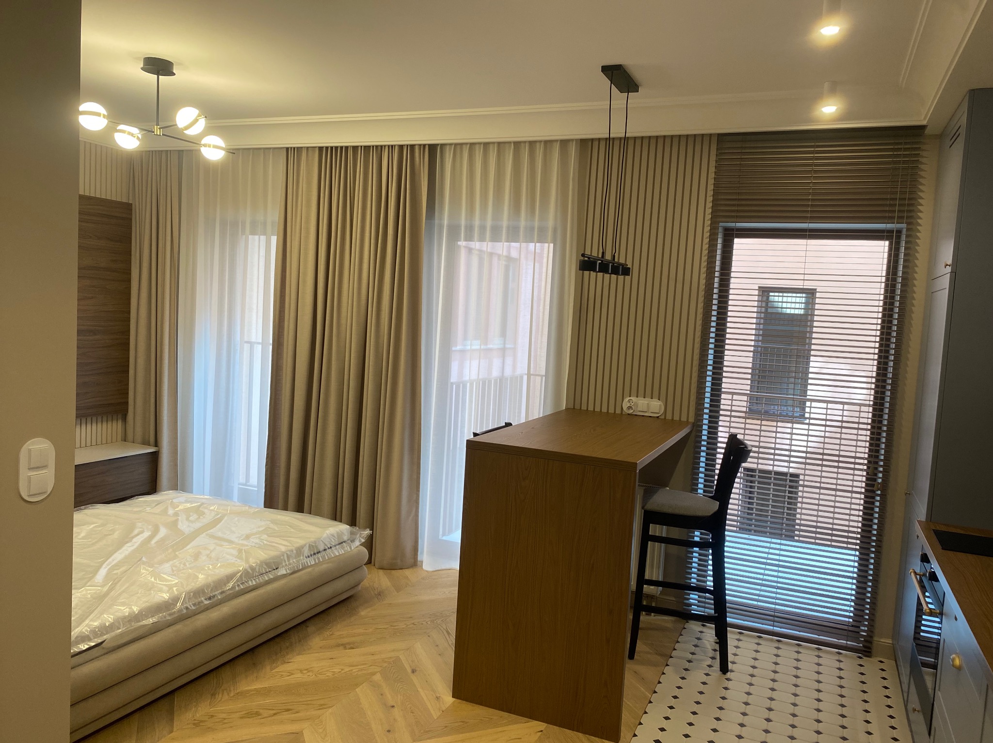Nowoczesne wnętrze: łóżko z folią ochronną, drewniany parkiet w jodełkę, barek z krzesłem, żaluzje i dekoracyjne lampy sufitowe. Neutralna kolorystyka.