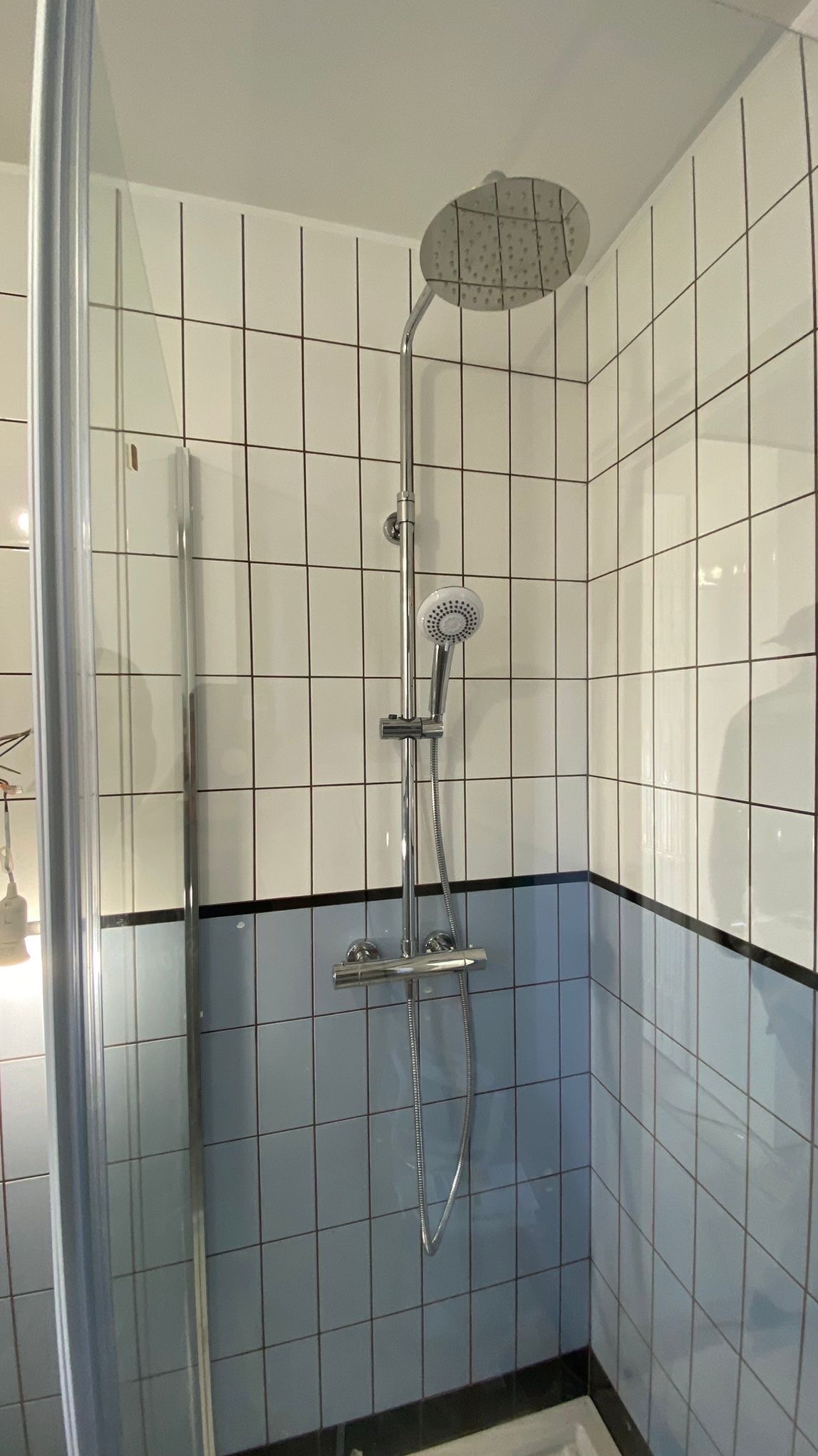 Nowoczesna kabina prysznicowa z chromowaną armaturą i biało-błękitnymi płytkami w kratkę. Widoczna szklana ściana kabiny i deszczownica.