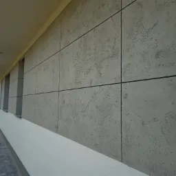 Elewacja domu jednorodzinnego z szarą, strukturalną powierzchnią imitującą beton, podzielona czarnymi fugami na prostokątne panele. Widoczne okna z białymi parapetami i fragment chodnika z kostki...