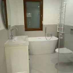 Nowoczesna łazienka z owalnym lustrem, białą szafką z umywalką, wanną i grzejnikiem drabinkowym, wykończona w jasnych odcieniach szarości i bieli.