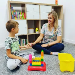 Kobieta w bluzie w jednorożce siedzi na podłodze z chłopcem, bawiąc się kolorowymi klockami na tle regału z książkami i zabawkami.