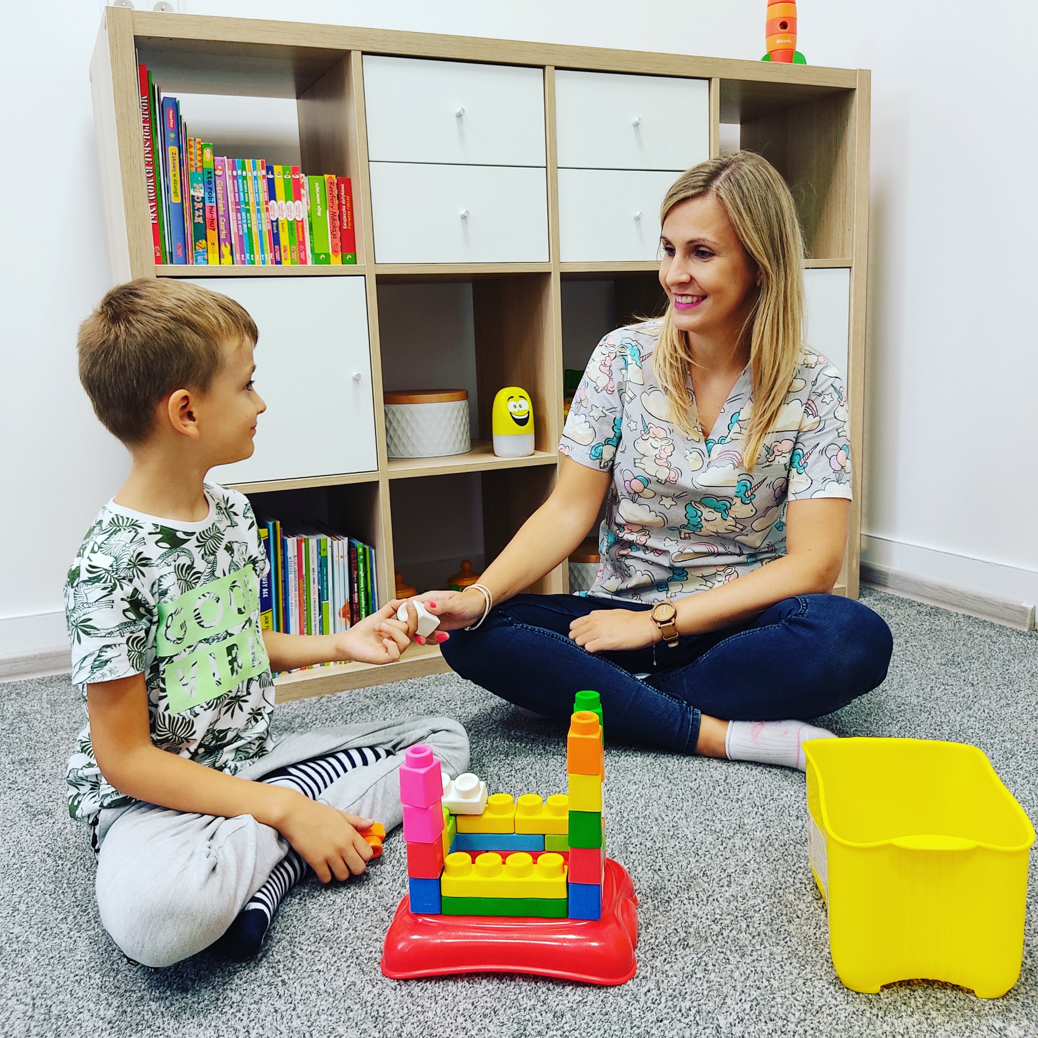 Kobieta w bluzie w jednorożce siedzi na podłodze z chłopcem, bawiąc się kolorowymi klockami na tle regału z książkami i zabawkami.
