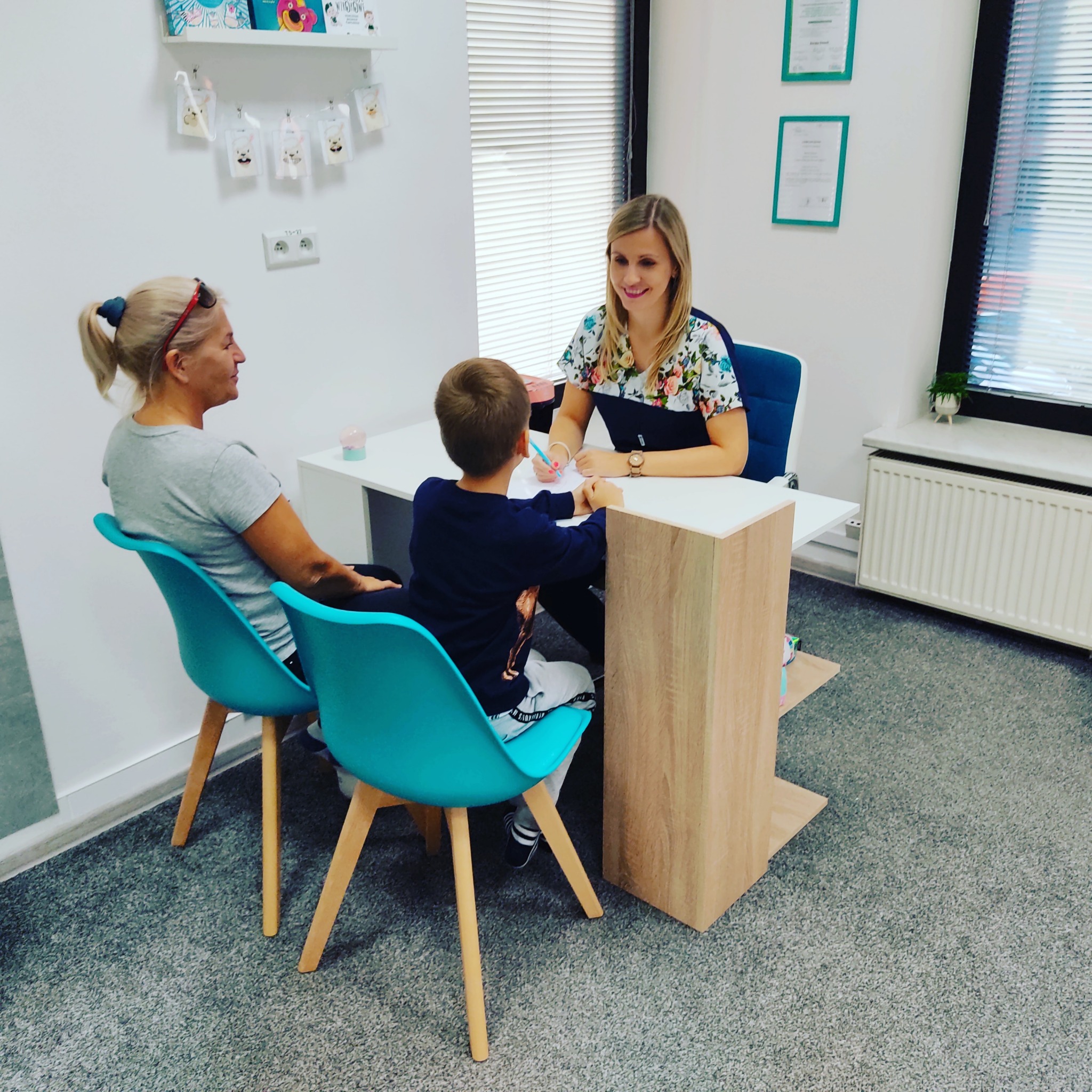 Kobieta w kwiecistej bluzce siedzi za biurkiem, uśmiechając się do dziecka rysującego na kartce, obok siedzi matka dziecka na turkusowym krześle.