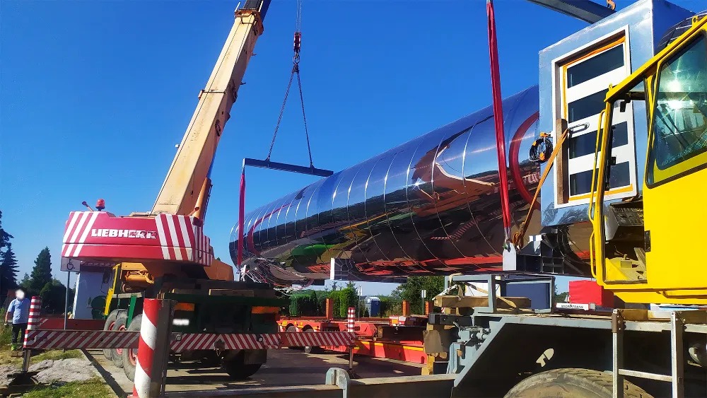 Żuraw Liebherr podnosi lśniącą, cylindryczną konstrukcję na tle błękitnego nieba. Widoczny fragment kabiny żurawia i platforma transportowa. Praca w toku.