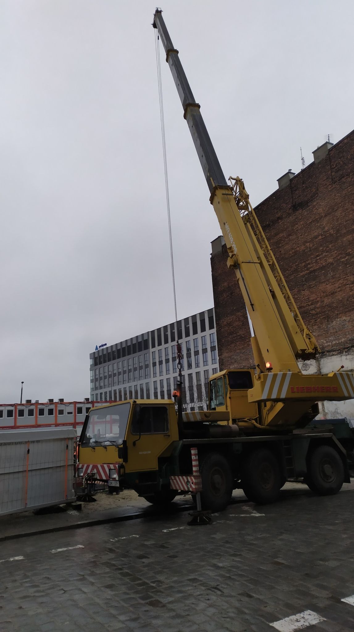 Żółty dźwig Liebherr na tle budynku biurowego i ceglanej ściany. Widoczne detale: tablice rejestracyjne, oznaczenia ostrzegawcze. Pochmurne niebo.