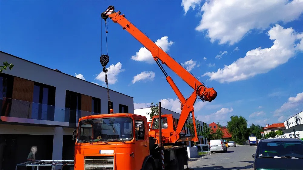 Pomarańczowy dźwig samochodowy z wysięgnikiem na tle błękitnego nieba i nowoczesnego budynku. Widoczne elementy konstrukcyjne i otoczenie placu budowy.