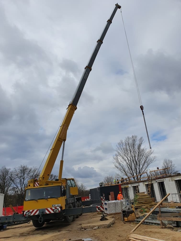 Żółty dźwig samochodowy na placu budowy, podnosi materiały budowlane. W tle widać budynek w trakcie wznoszenia i pochmurne niebo.
