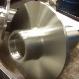 Dwa metalowe elementy obrotowe z kołnierzem leżące na brudnym metalowym stole warsztatowym, widoczne ślady obróbki skrawaniem.