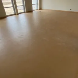 Jasnobrązowa, jednolita powierzchnia podłogi w przestronnym, jasnym pomieszczeniu z trzema oknami; widoczne ślady stóp na podłodze i surowy sufit podwieszany.