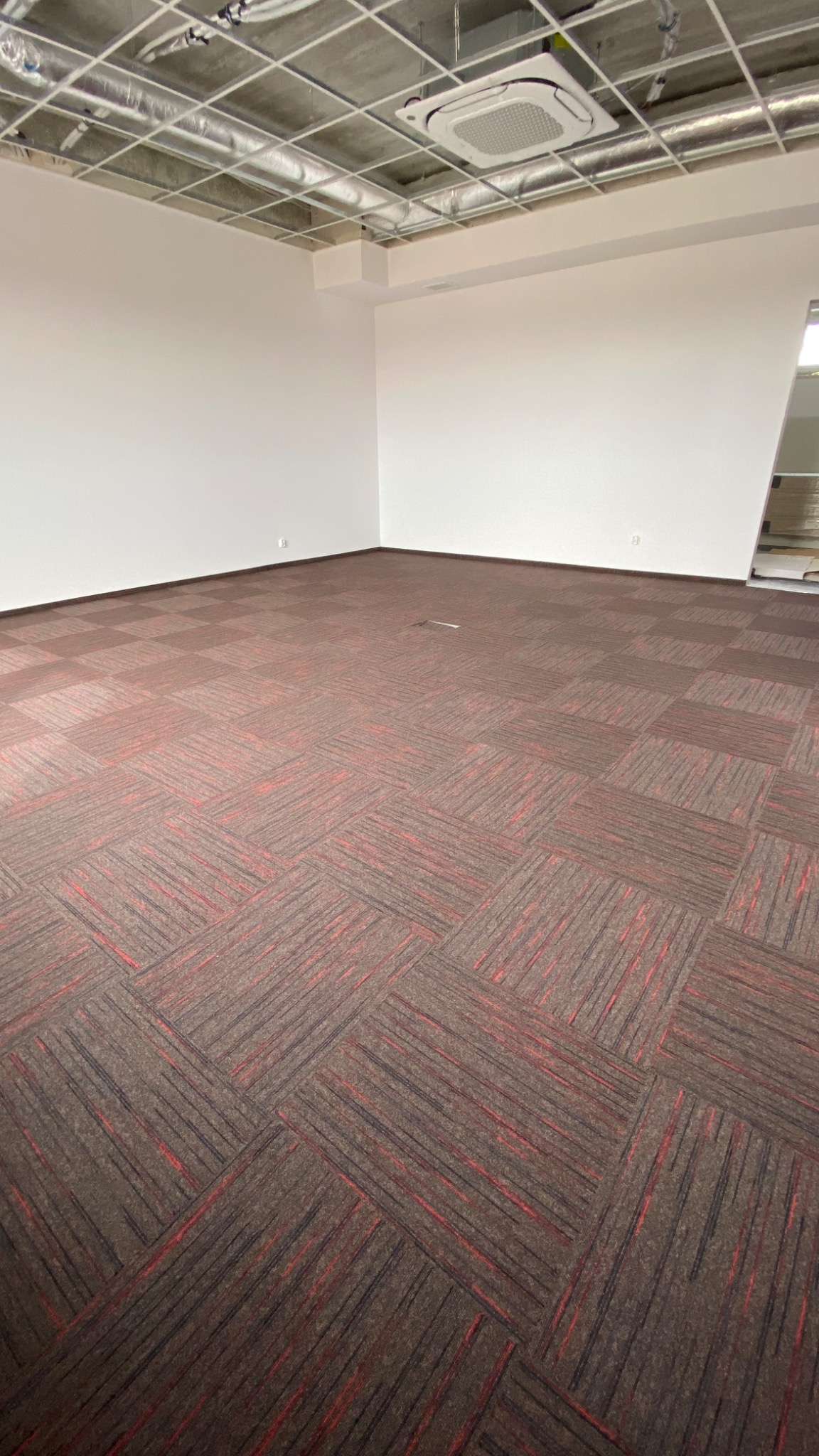Wykładzina dywanowa w brązowo-czerwone kwadraty w pustym biurze z białą ścianą i widoczną instalacją sufitową.
