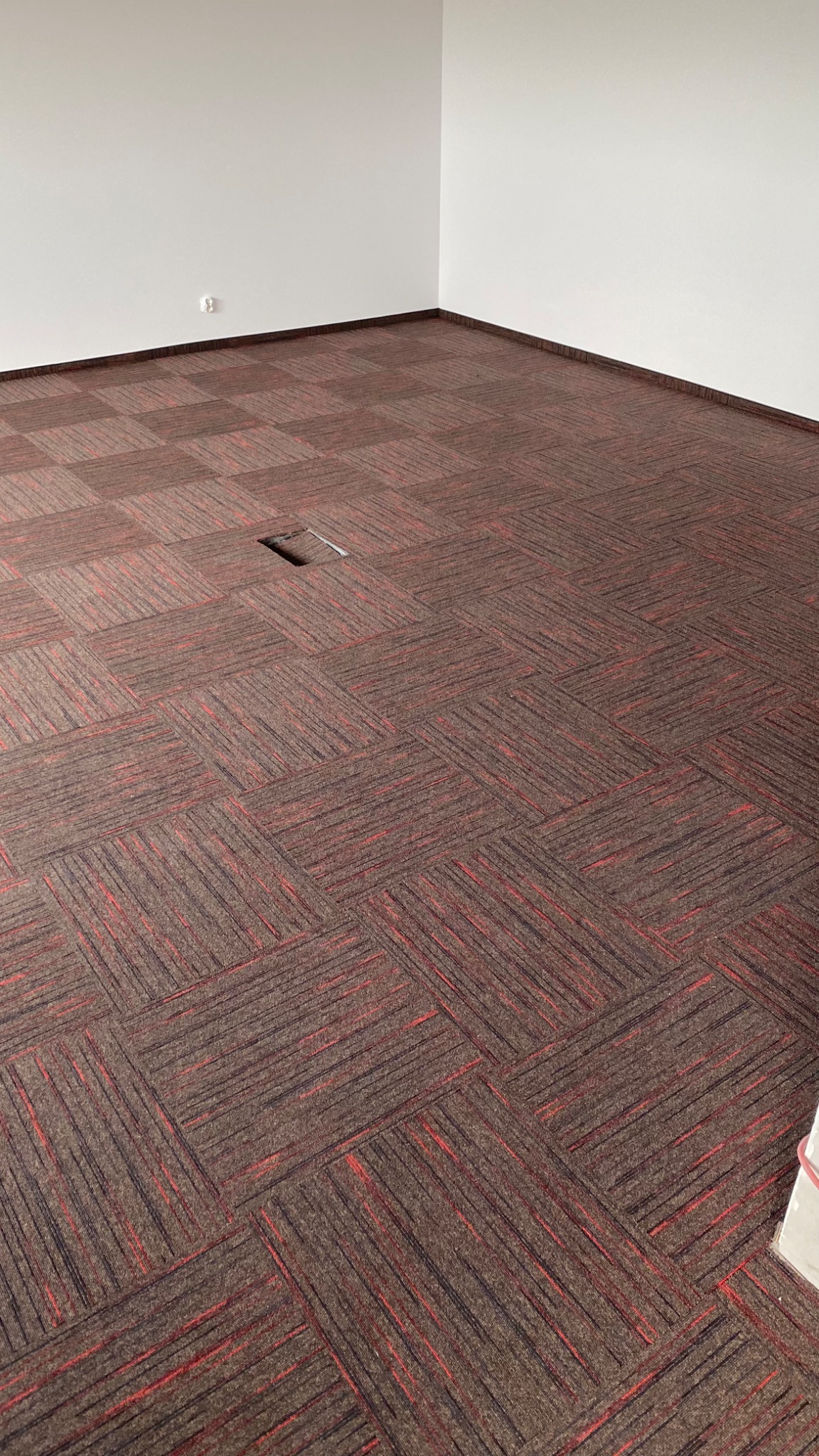 Wykładzina dywanowa w brązowo-czerwony wzór ułożona w kwadraty, widoczny fragment podłogi z gniazdem elektrycznym w nowym biurze.