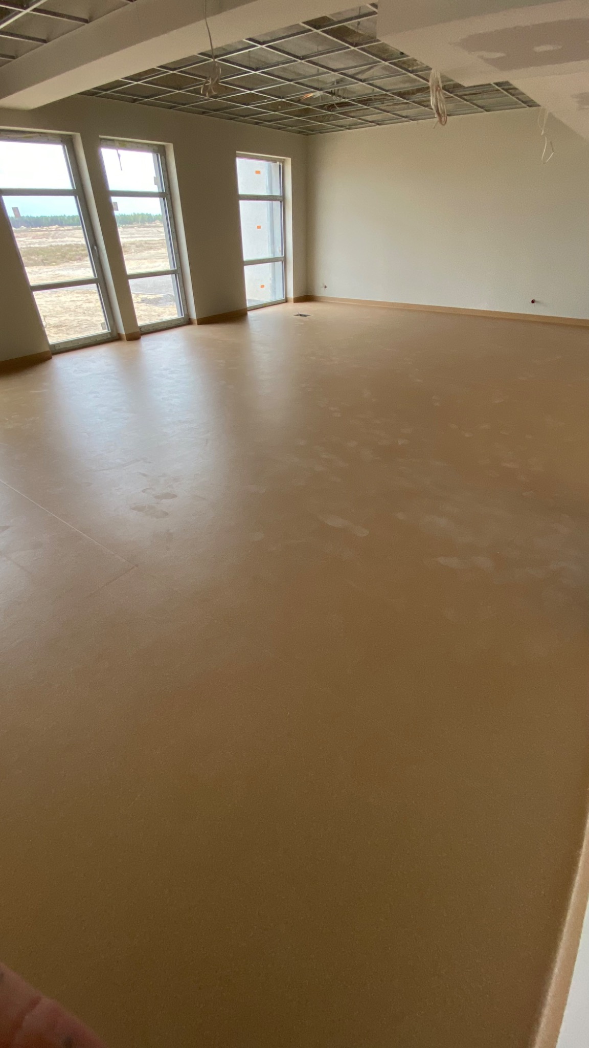 Jasnobrązowa, jednolita powierzchnia podłogi w przestronnym, jasnym pomieszczeniu z trzema oknami; widoczne ślady stóp na podłodze i surowy sufit podwieszany.