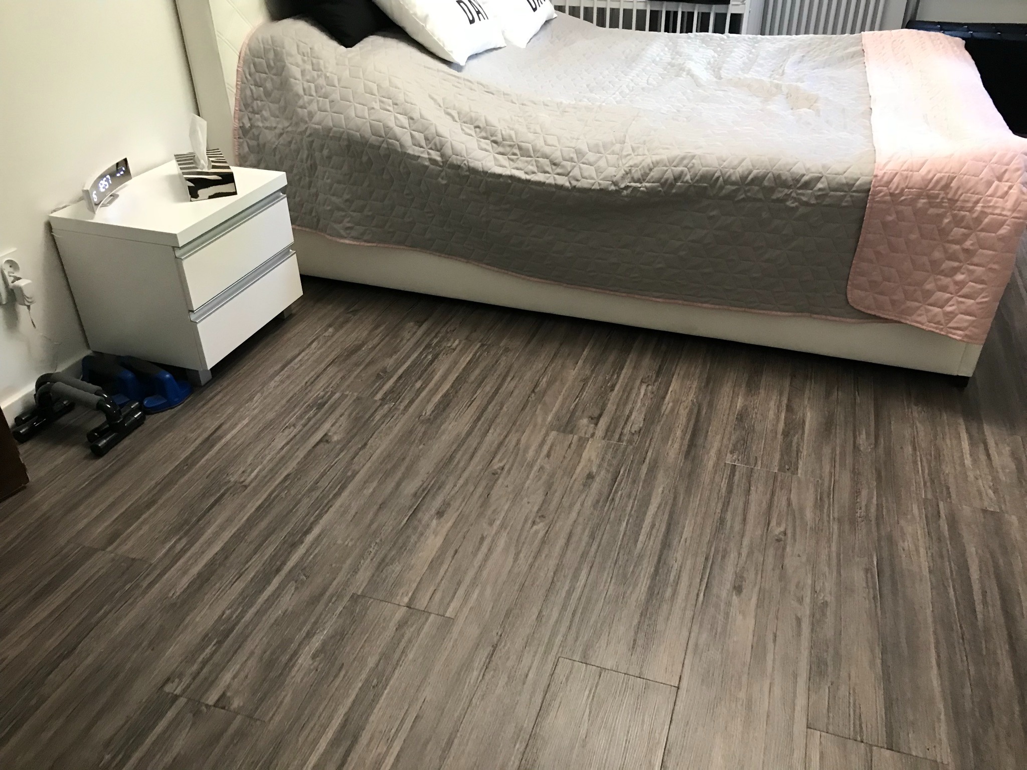 Szare panele podłogowe ułożone w sypialni, widoczne przy łóżku i szafce nocnej z akcesoriami do ćwiczeń w tle.