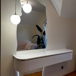 B.art Pracownia Stolarska Bartłomiej Kijowski - Biała toaletka z lustrem o nieregularnym kształcie, podświetlana od dołu. W lustrze odbija się sofa i roślina doniczkowa. Nowoczesny design.