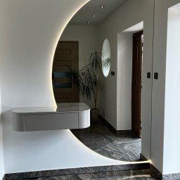 B.art Pracownia Stolarska Bartłomiej Kijowski - Nowoczesny przedpokój z podświetlanym lustrem w kształcie półksiężyca i wiszącą szafką. Na podłodze szara, wzorzysta posadzka. W tle widoczna roślina doniczkowa.