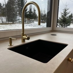 B.art Pracownia Stolarska Bartłomiej Kijowski - Nowoczesny blat kuchenny z czarnym zlewem i złotą baterią na tle okna z widokiem na zimowy krajobraz. Minimalistyczny design, jasne kolory, naturalne światło.