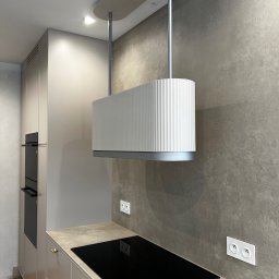 B.art Pracownia Stolarska Bartłomiej Kijowski - Nowoczesna kuchnia z beżowymi szafkami, czarną płytą indukcyjną i designerskim, białym okapem z żłobieniami, zawieszonym na srebrnych rurach.