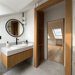 B.art Pracownia Stolarska Bartłomiej Kijowski - Nowoczesna łazienka na poddaszu z drewnianą szafką pod umywalką, okrągłym lustrem z podświetleniem i drewnianą ramą drzwiową z lamelami. Widok na pokój z oknem dachowym.