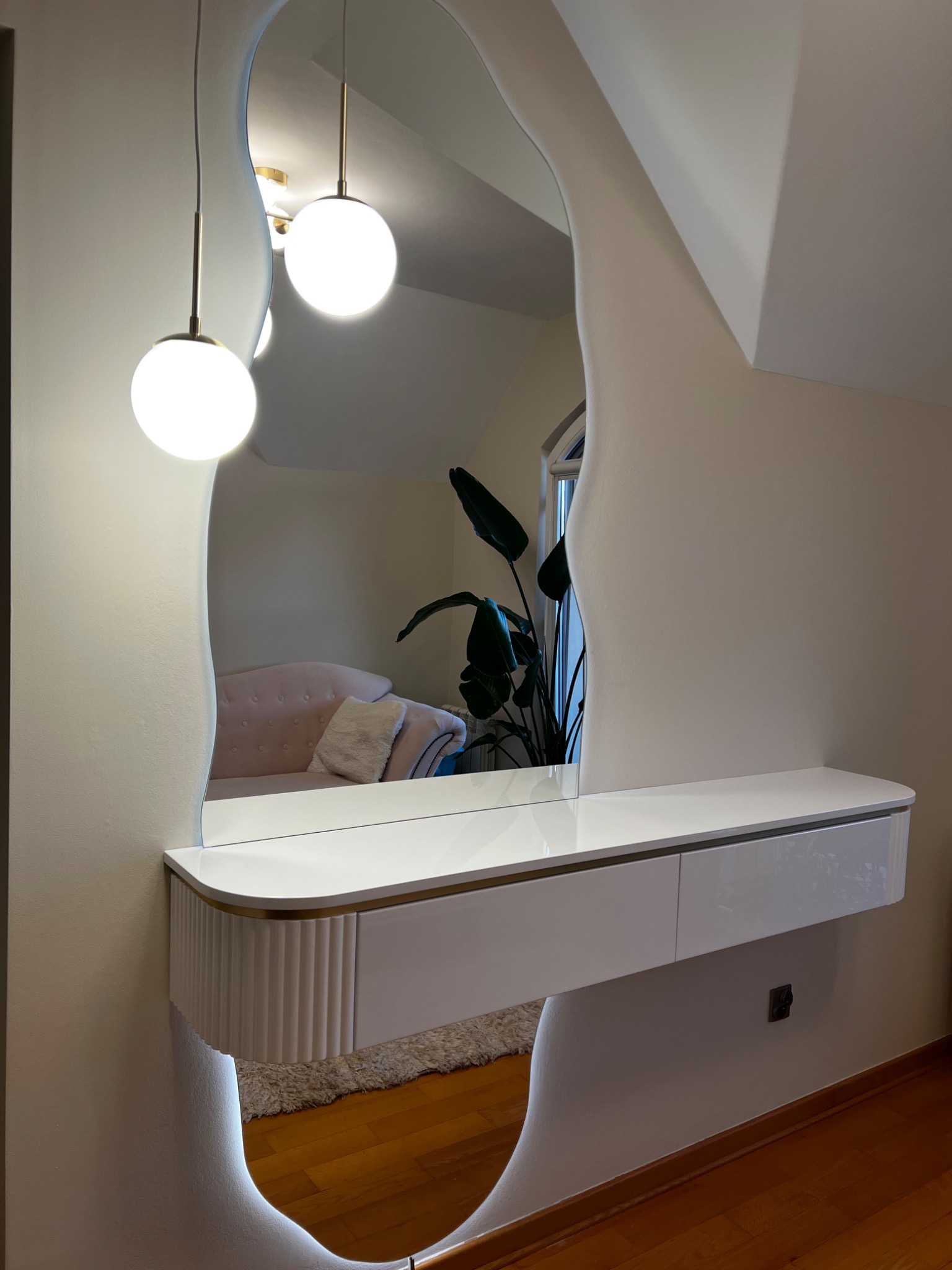 Biała toaletka z lustrem o nieregularnym kształcie, podświetlana od dołu. W lustrze odbija się sofa i roślina doniczkowa. Nowoczesny design.
