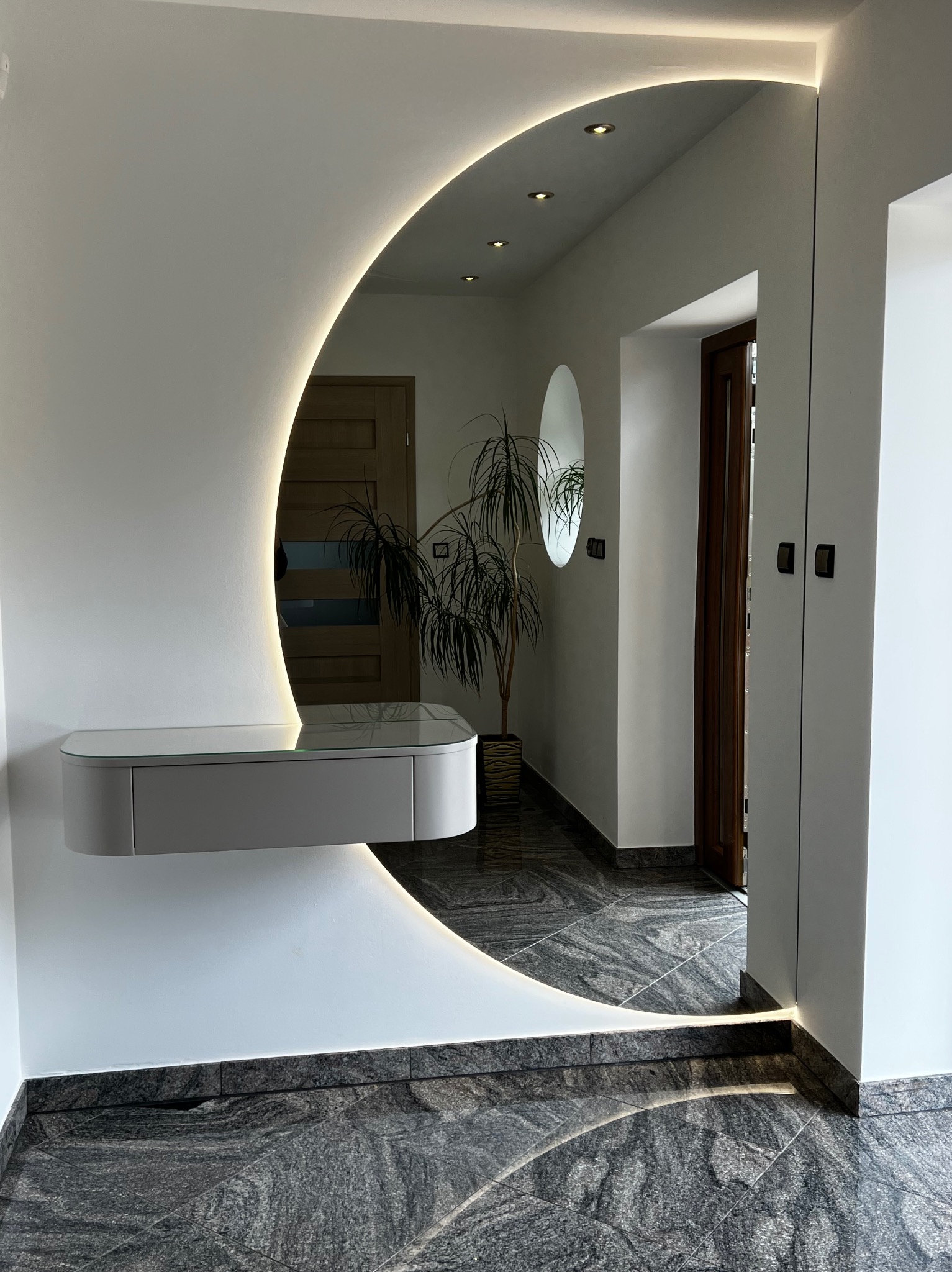 Nowoczesny przedpokój z podświetlanym lustrem w kształcie półksiężyca i wiszącą szafką. Na podłodze szara, wzorzysta posadzka. W tle widoczna roślina doniczkowa.