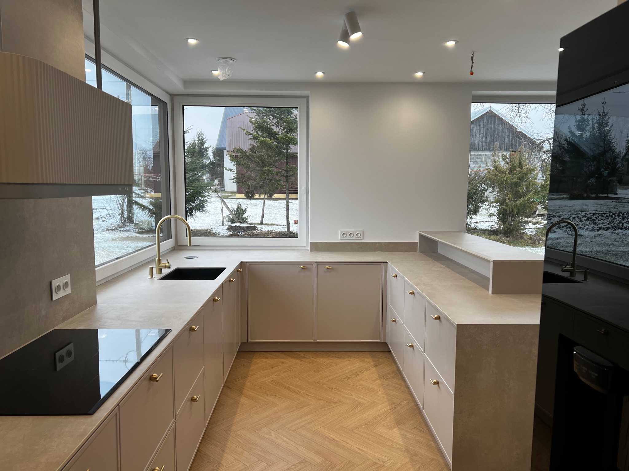 Jasna kuchnia z drewnianą podłogą w jodełkę, złote detale armatury i uchwytów, duże okna z widokiem na zimowy krajobraz. Minimalistyczny design.