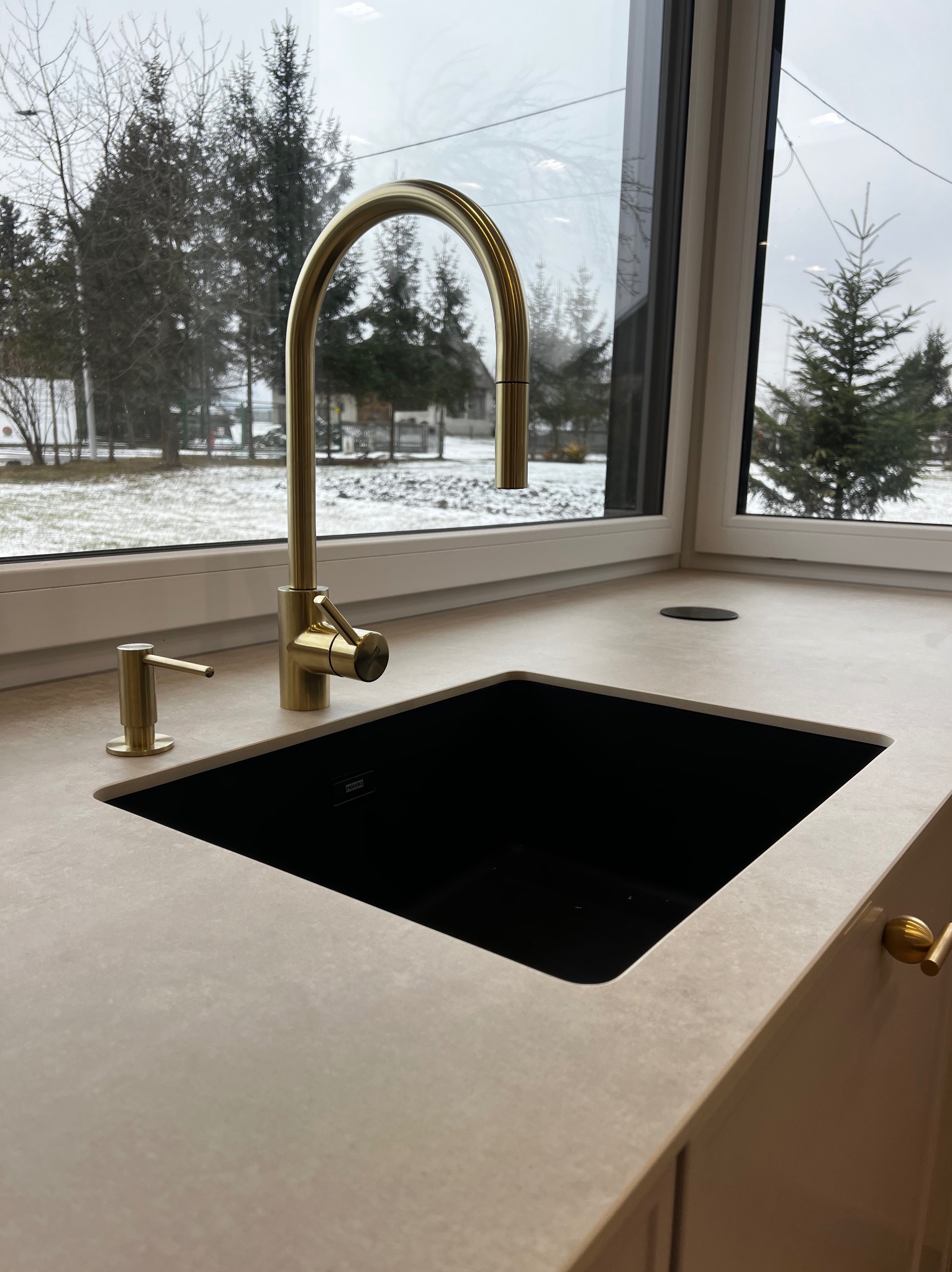 Nowoczesny blat kuchenny z czarnym zlewem i złotą baterią na tle okna z widokiem na zimowy krajobraz. Minimalistyczny design, jasne kolory, naturalne światło.