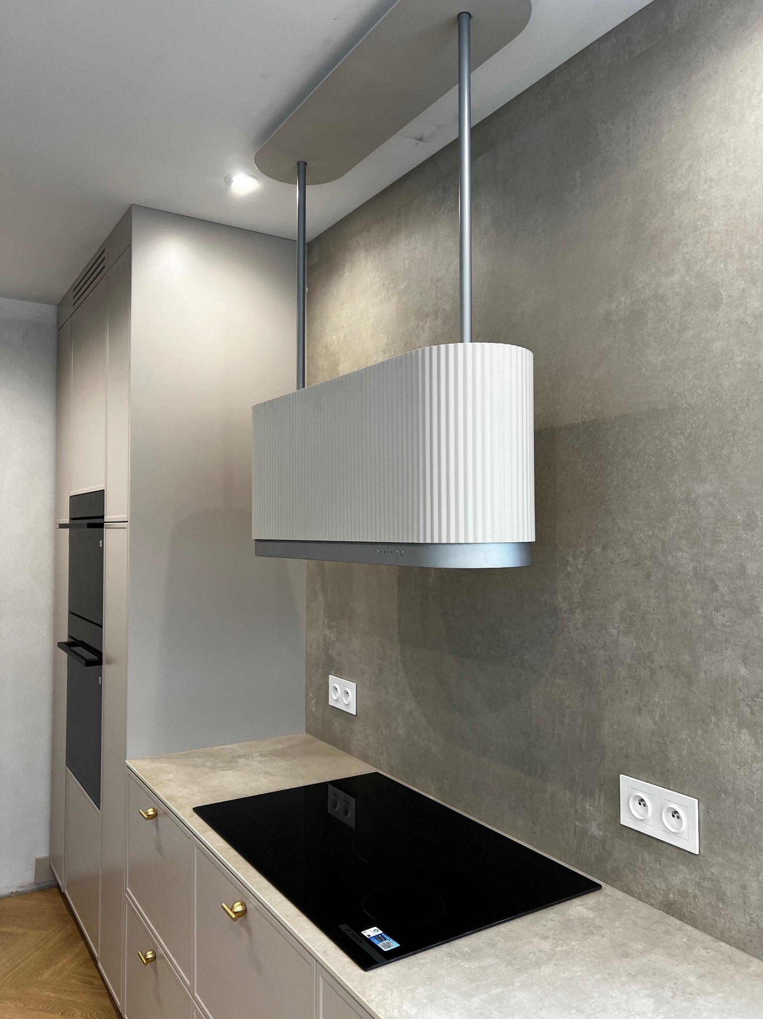 Nowoczesna kuchnia z beżowymi szafkami, czarną płytą indukcyjną i designerskim, białym okapem z żłobieniami, zawieszonym na srebrnych rurach.