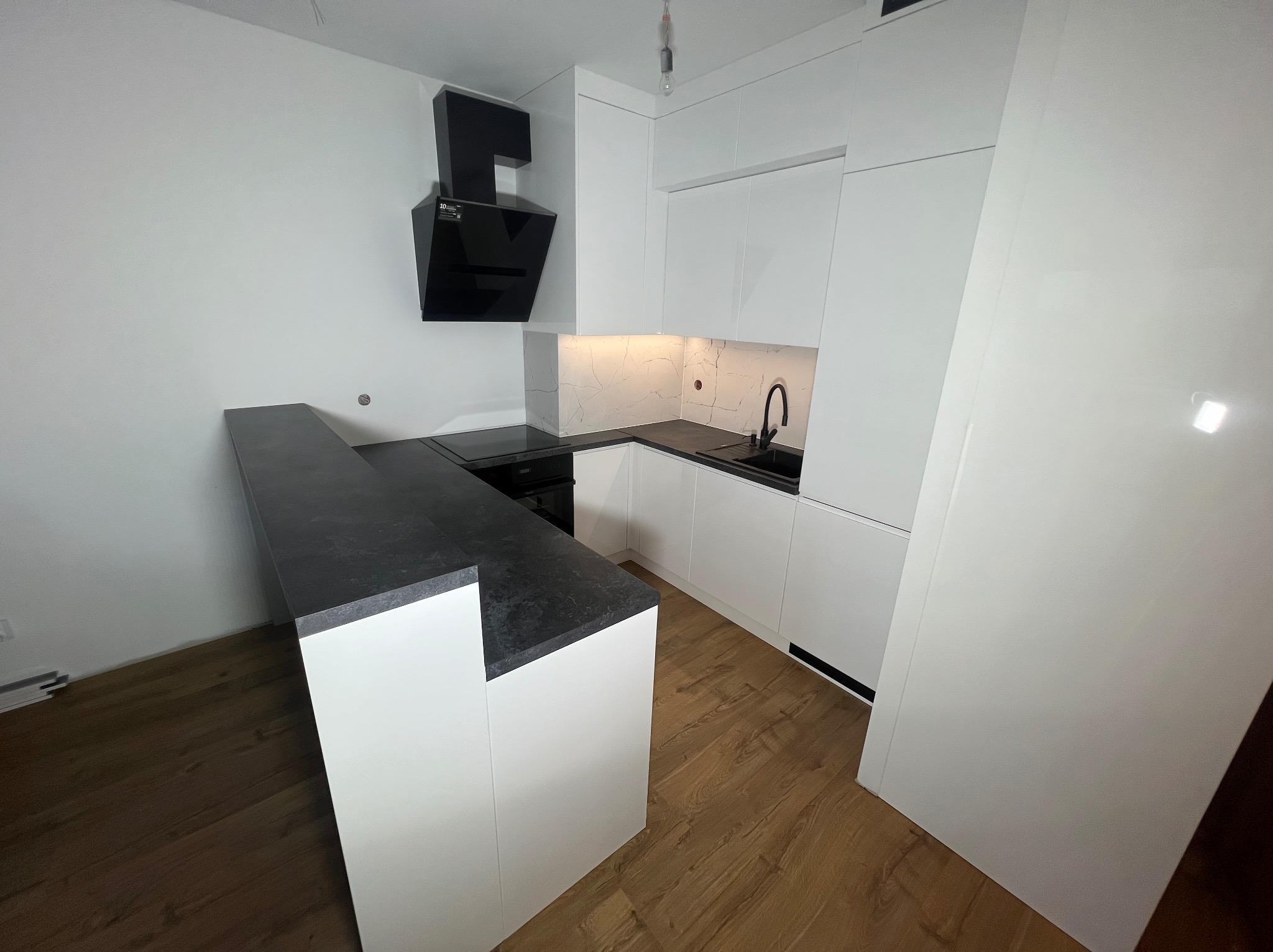 Nowoczesna, biała kuchnia z czarnym blatem i okapem w Krosno. Minimalistyczny design, marmurowe wykończenie ściany nad blatem, drewniana podłoga.