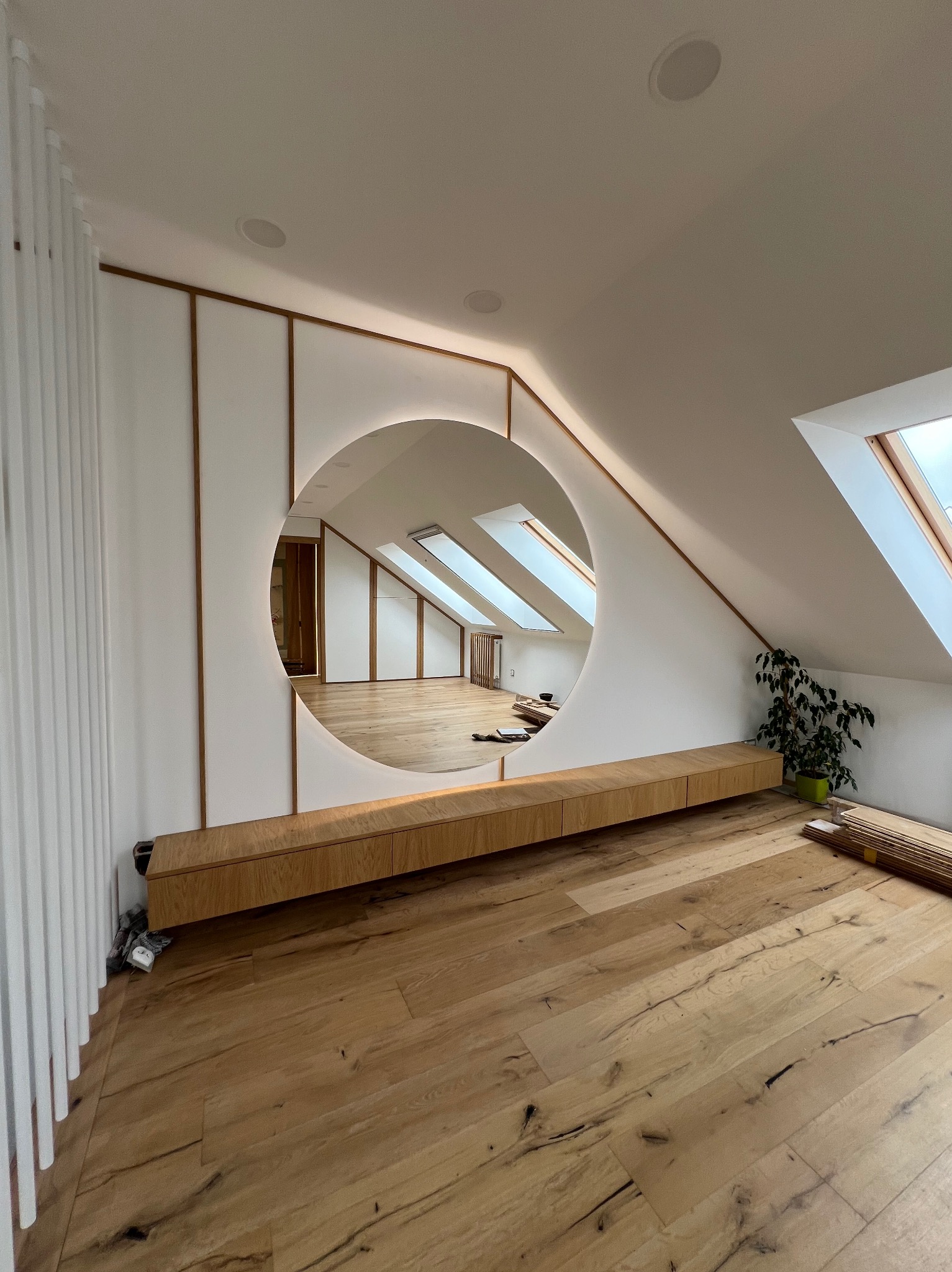 Stylowa szafka RTV z drewna wkomponowana w ścianę z okrągłym, podświetlanym otworem, widok na poddasze z oknami dachowymi i drewnianą podłogą.