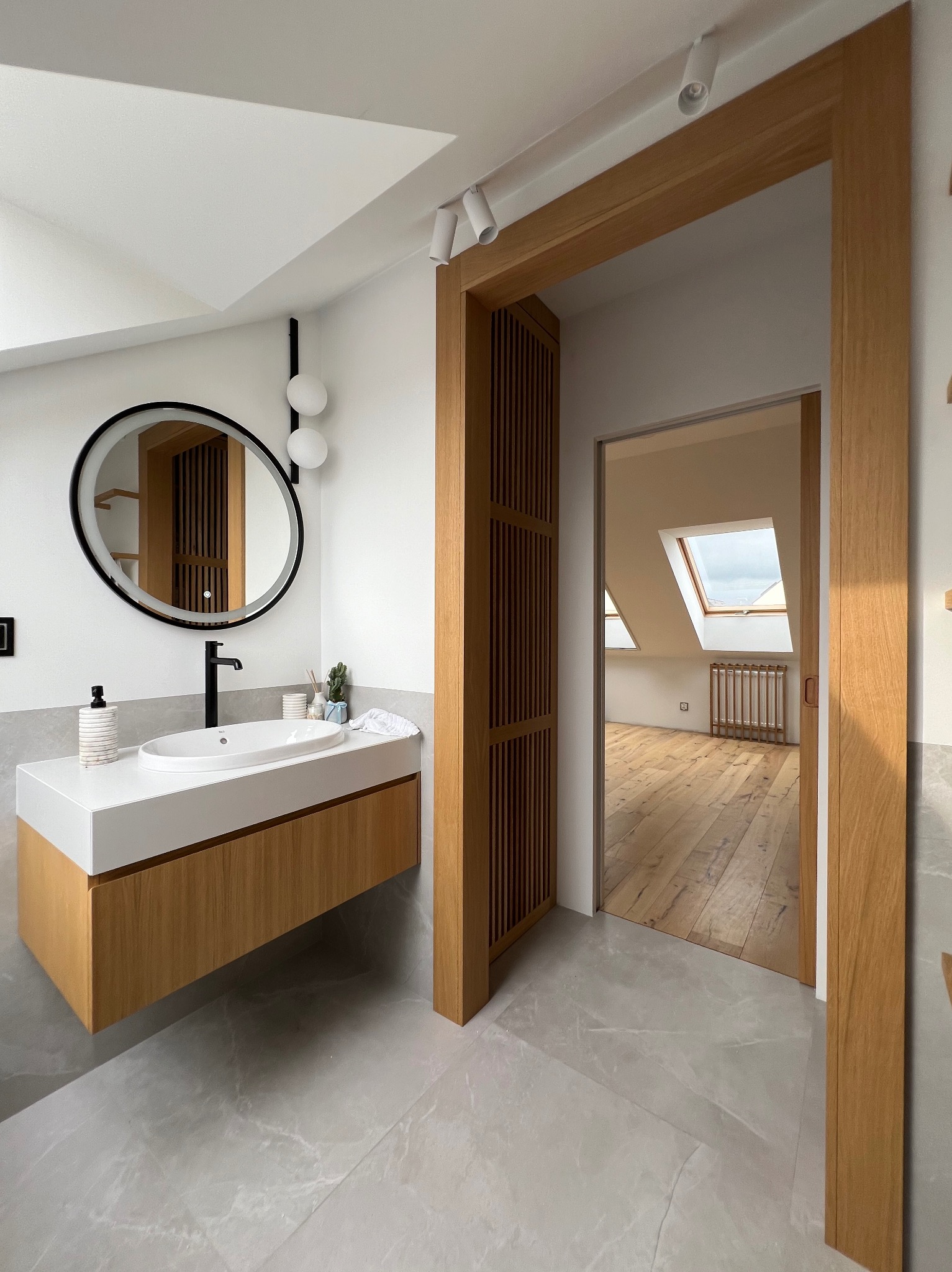 Nowoczesna łazienka na poddaszu z drewnianą szafką pod umywalką, okrągłym lustrem z podświetleniem i drewnianą ramą drzwiową z lamelami. Widok na pokój z oknem dachowym.