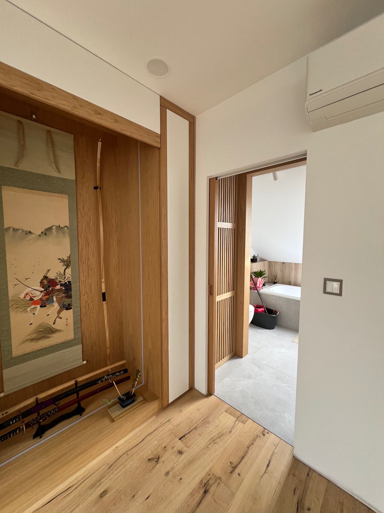 Nowoczesne wnętrze z drewnianą zabudową w stylu japońskim: przesuwne drzwi, dekoracyjna wnęka z katanami i obrazem, widok na łazienkę. Jasne kolory, minimalistyczny design.